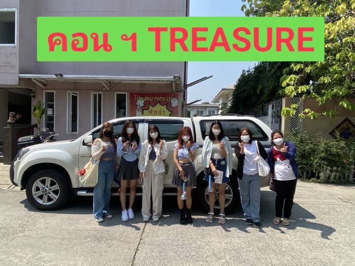 A group of six individuals stands in front of a large white vehicle parked on a well-maintained outdoor area. They are wearing casual clothing and masks, creating a friendly and approachable atmosphere. A green sign with the text 'TREASURE' is prominently displayed above.