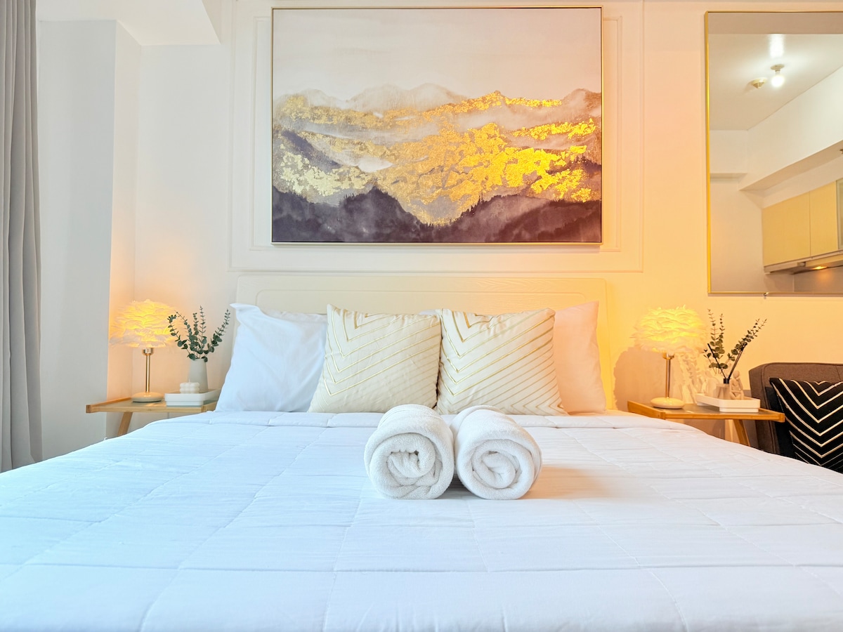 A bright and inviting sleeping area features a spacious bed adorned with white linens. Two decorative pillows sit at the head of the bed, complemented by a golden-framed artwork above. Soft lighting is provided by bedside lamps, illuminating the tranquil atmosphere.