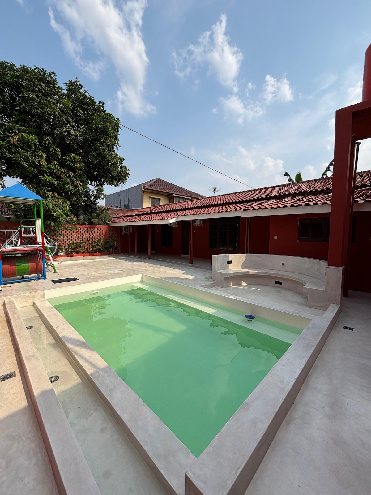 A private swimming pool is nestled within a spacious courtyard. Surrounding the pool, cream-colored stone steps provide seating and a smooth entry. Lush greenery and a vibrant red exterior complement the serene environment, while blue skies and scattered clouds add to the outdoor ambiance.