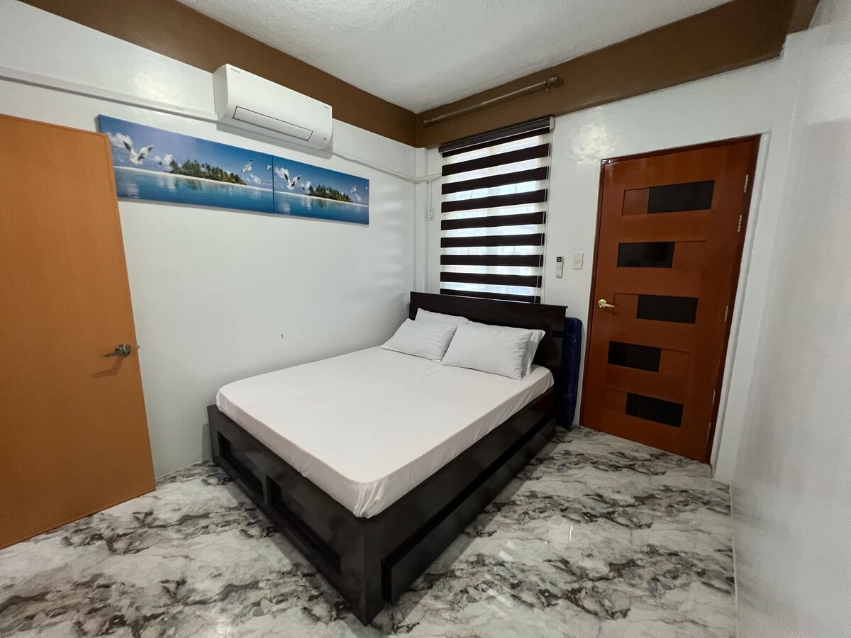 A well-lit sleeping area features a spacious bed with neatly arranged pillows. Above the bed, artwork depicting a serene landscape is displayed. Dark striped curtains frame a window, while modern doors and a light-colored floor contribute to a clean, uncluttered aesthetic.