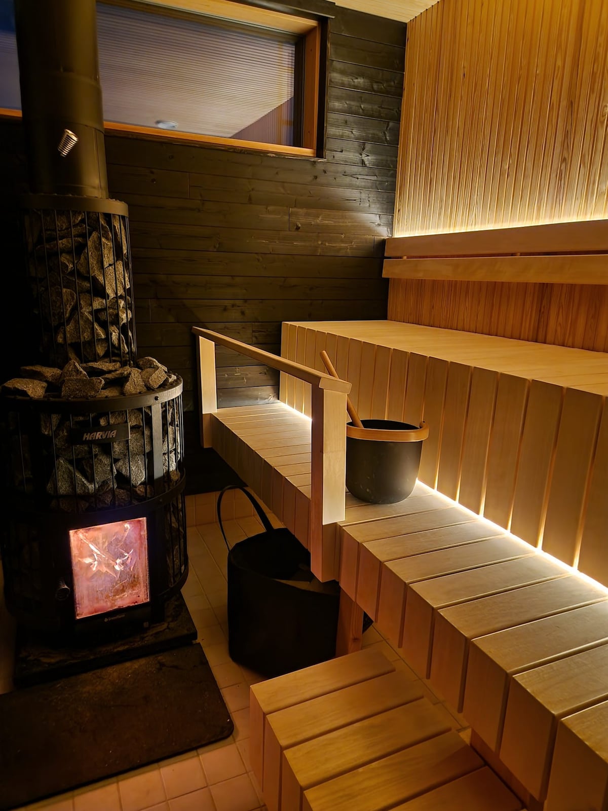A wood-paneled sauna features tiered wooden seating and ambient lighting. A traditional wood-burning stove is positioned in the corner, with a bucket and ladle nearby. The warm glow from the stove enhances the tranquil atmosphere, inviting relaxation.