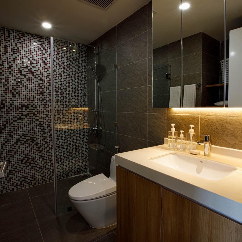A modern bathroom features a glass-enclosed shower with a mosaic tile backdrop. The countertop has a sleek design and is equipped with toiletries. Soft lighting highlights the space, while two sets of neatly arranged towels are visible on a shelf.