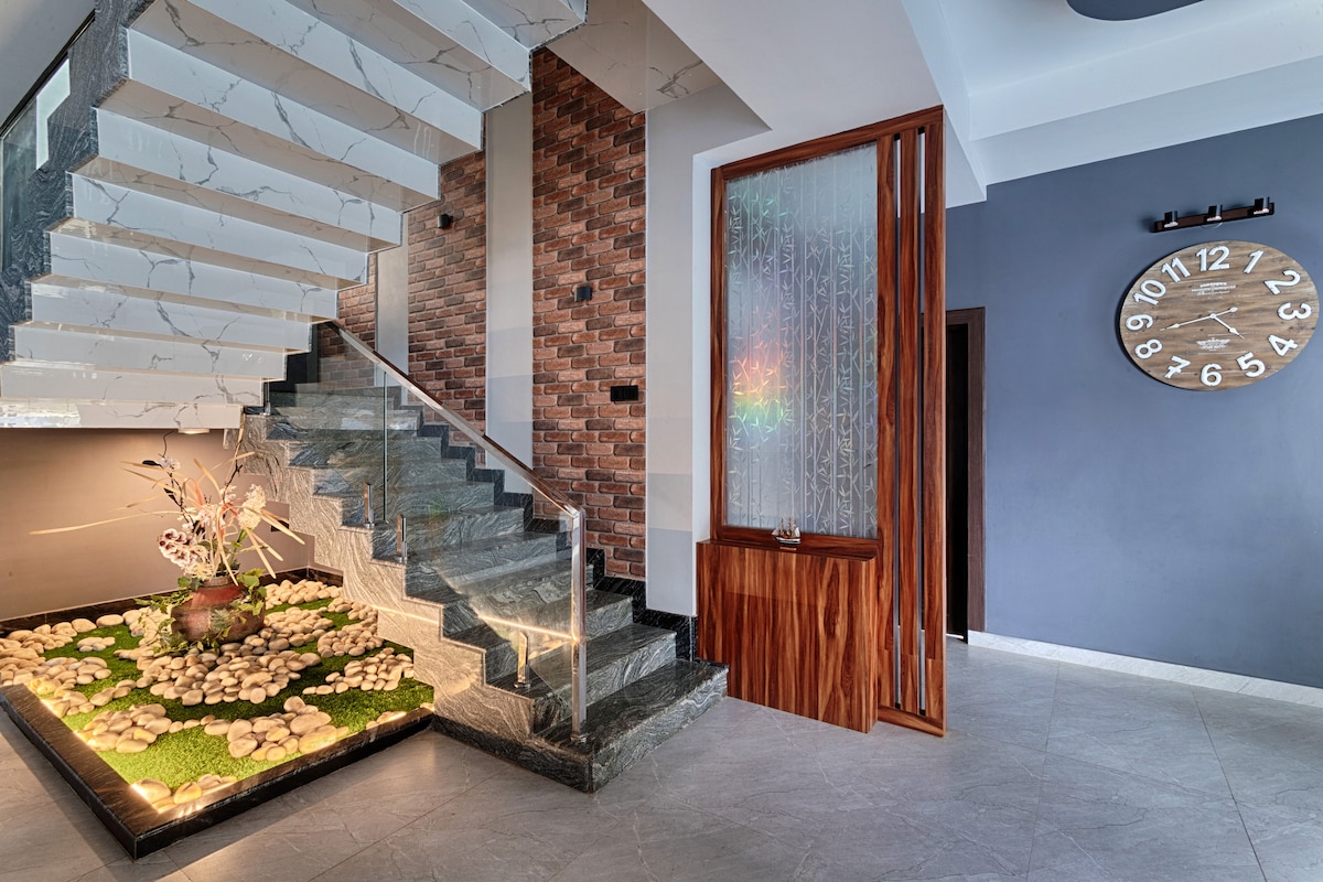 An inviting entrance area features a modern staircase with a glass railing and elegantly designed steps. Natural stones and a decorative arrangement of plants accentuate the lower level, while a large wall clock adds a contemporary touch to the aesthetic.
