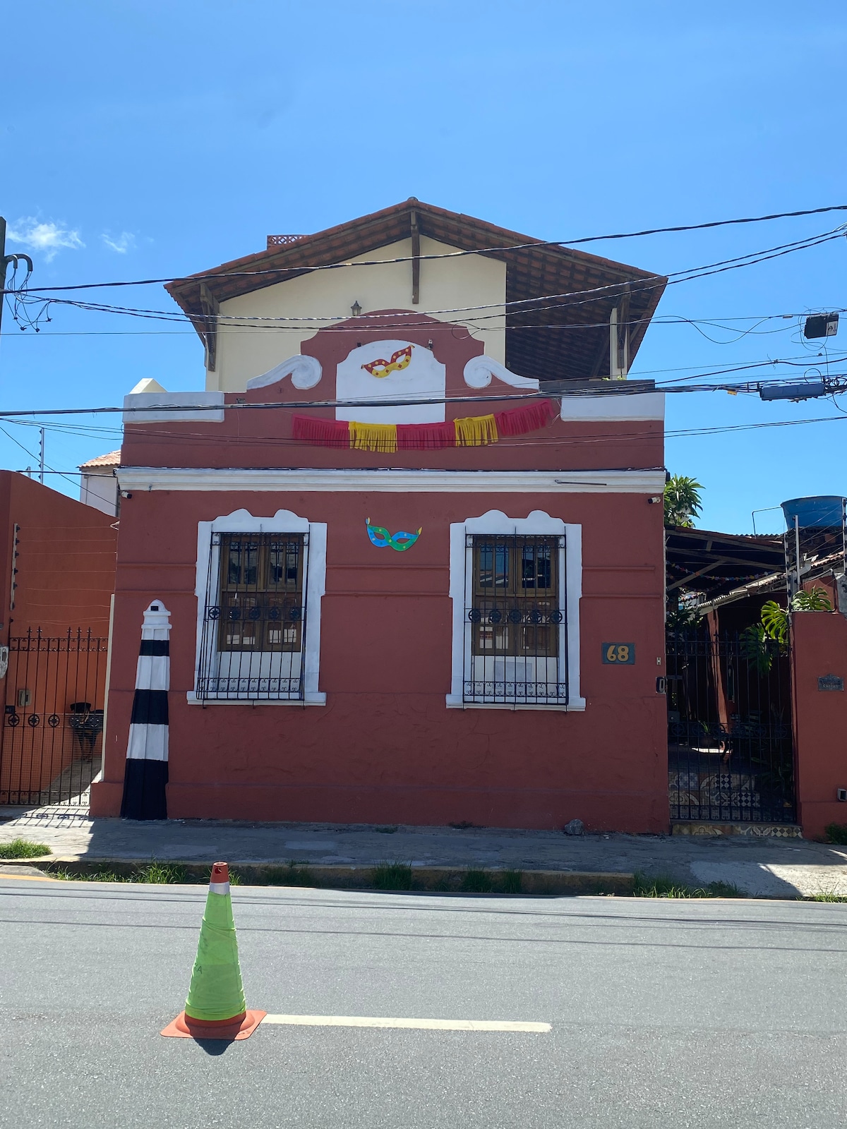 A charming two-story building is displayed in a vibrant coral color, with white trim around the windows and doors. Colorful festive decorations hang above a decorative emblem. A small gate offers access to the property, framed by a simple white fence.