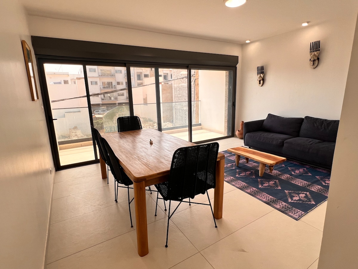 A minimalist living room features a dark sofa and a wooden coffee table on a patterned rug. A large dining table with black chairs is positioned near expansive glass doors, opening to a balcony that allows natural light to brighten the space.