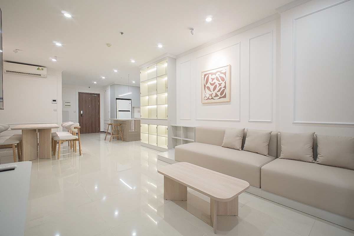 A spacious living area features a neutral color palette with soft seating. A minimalistic coffee table is positioned in front of a sectional sofa. Light-colored wooden chairs and a dining table are visible nearby, alongside built-in shelving that adds contemporary style.