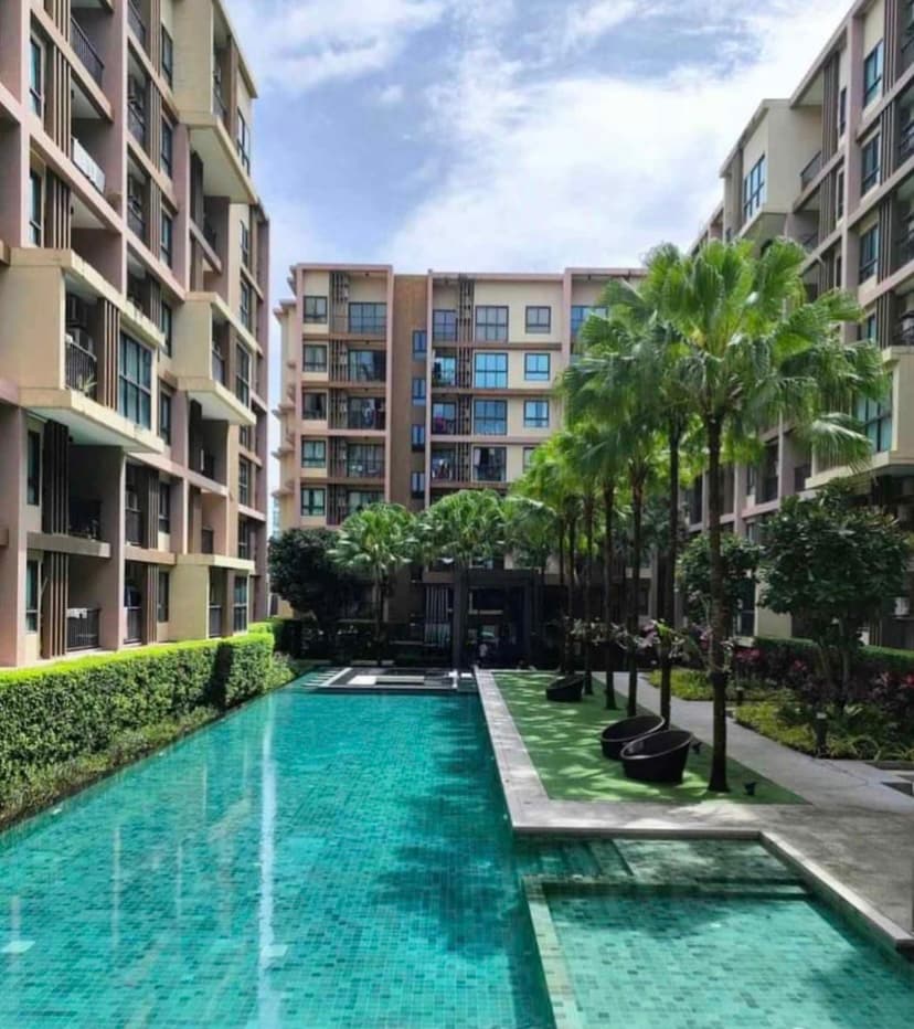 The image showcases a tranquil swimming pool, bordered by lush greenery and palm trees. Several lounge chairs are positioned along the pool’s edge, complementing the surrounding architecture of modern condominium buildings under a clear sky.