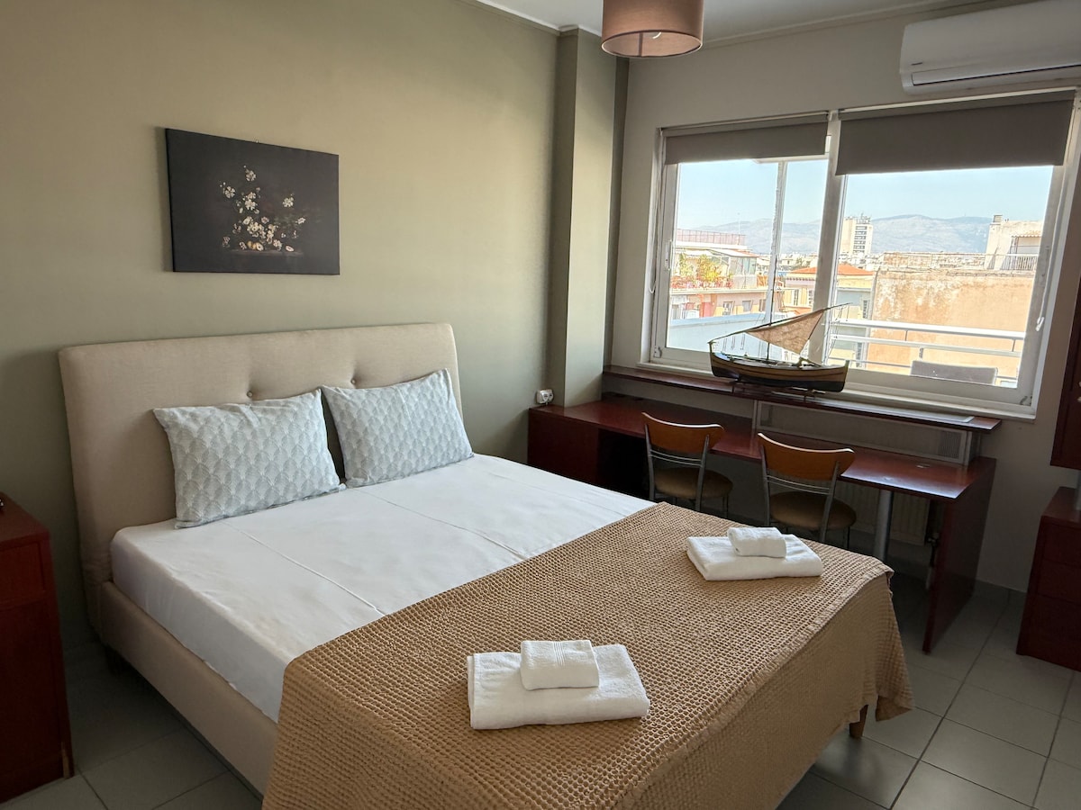 A comfortable bedroom is seen with a spacious bed featuring light-colored linens and two decorative pillows. A minimalist artwork hangs on the green wall above the bed. A small desk with two chairs is positioned by a window overlooking the cityscape.