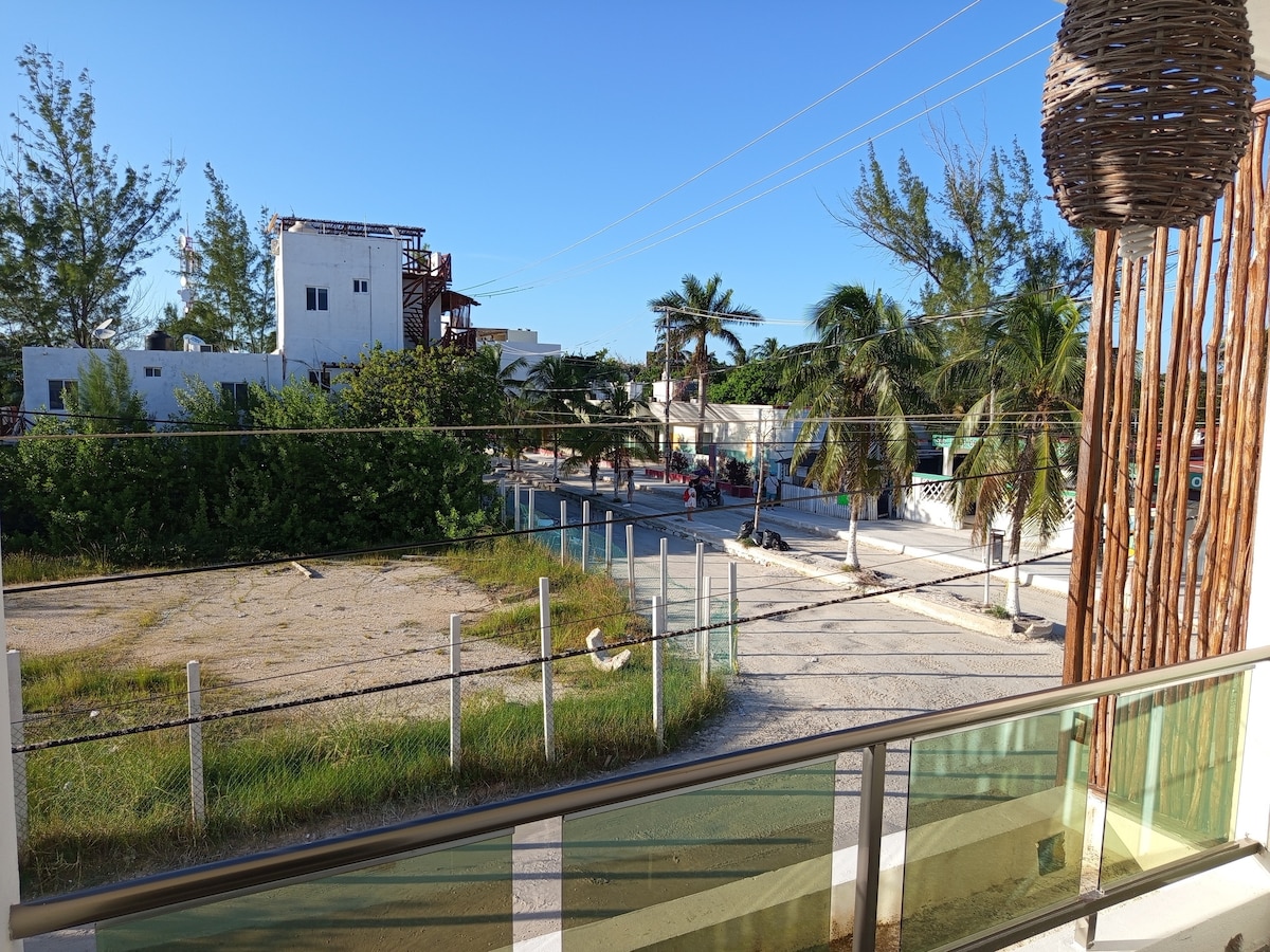 A view of the surroundings from a balcony showcases a quiet street lined with palm trees and greenery. Sunlight illuminates the area, revealing both sandy patches and paved roads. Buildings in varying styles are visible in the distance, creating a sense of the local environment.