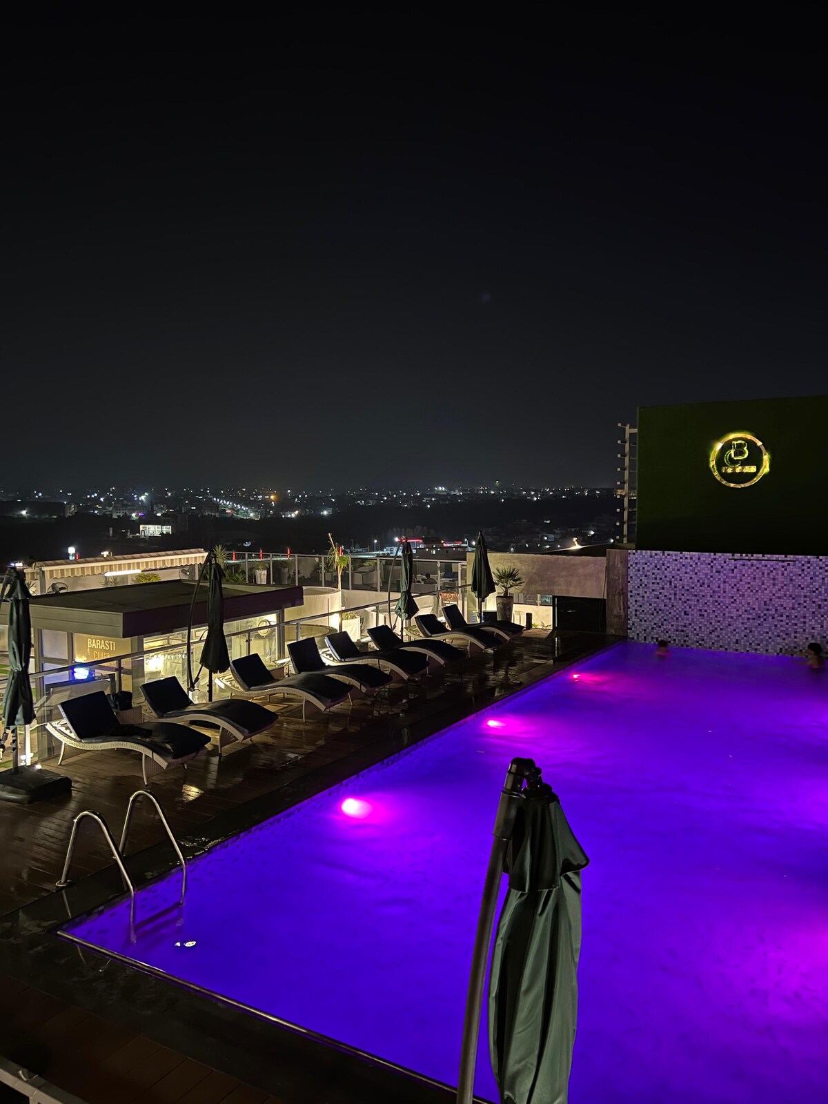 A rooftop pool area is illuminated with purple lighting, reflecting the night sky. Loungers are arranged around the pool, and umbrellas are positioned nearby. The city skyline is visible in the background, creating a serene atmosphere for evening relaxation.