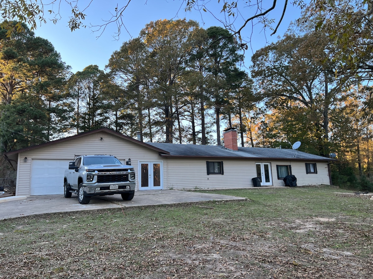 A single-story house is set on a spacious lawn, surrounded by tall trees. A gravel driveway leads to a garage, and a white truck is parked in front. The exterior features light-colored siding and a covered entryway with double doors.