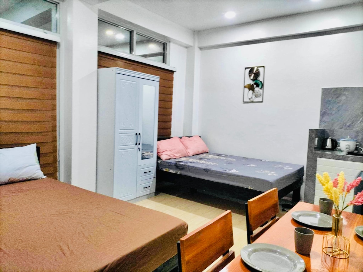 A studio space is presented, featuring two double beds adorned with linens in neutral and pastel tones. A wardrobe stands between the beds, and a dining table with four chairs occupies the area, complemented by a kitchenette in the background.