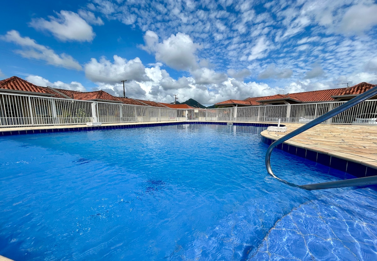A spacious pool is visible under a clear blue sky, surrounded by a well-maintained deck. The water's surface reflects the clouds above, creating a serene atmosphere. A partial view of surrounding buildings and fencing is seen in the background.