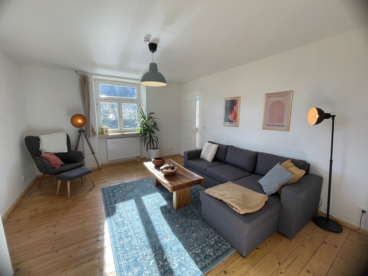 A living room setup features a gray sectional sofa and wooden coffee table on a patterned rug. A reading chair sits in one corner, complemented by a floor lamp. Natural light streams through a window adorned with light curtains, creating a cozy atmosphere.