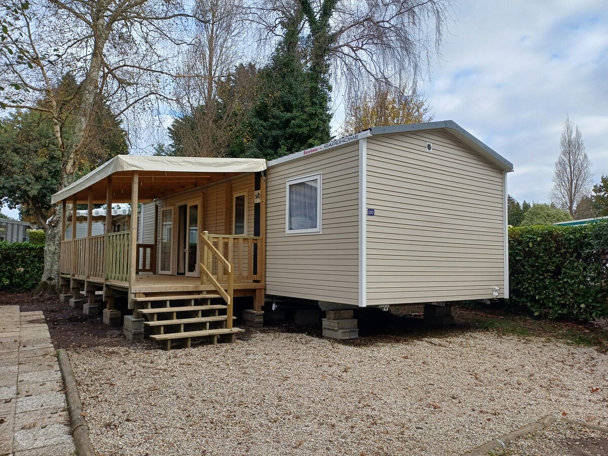 A mobile home is set within a green landscape, featuring a covered wooden terrace with steps leading to the entrance. The exterior is clad in light-colored siding, complemented by gravel pathways and surrounding foliage, offering a calm and inviting environment.