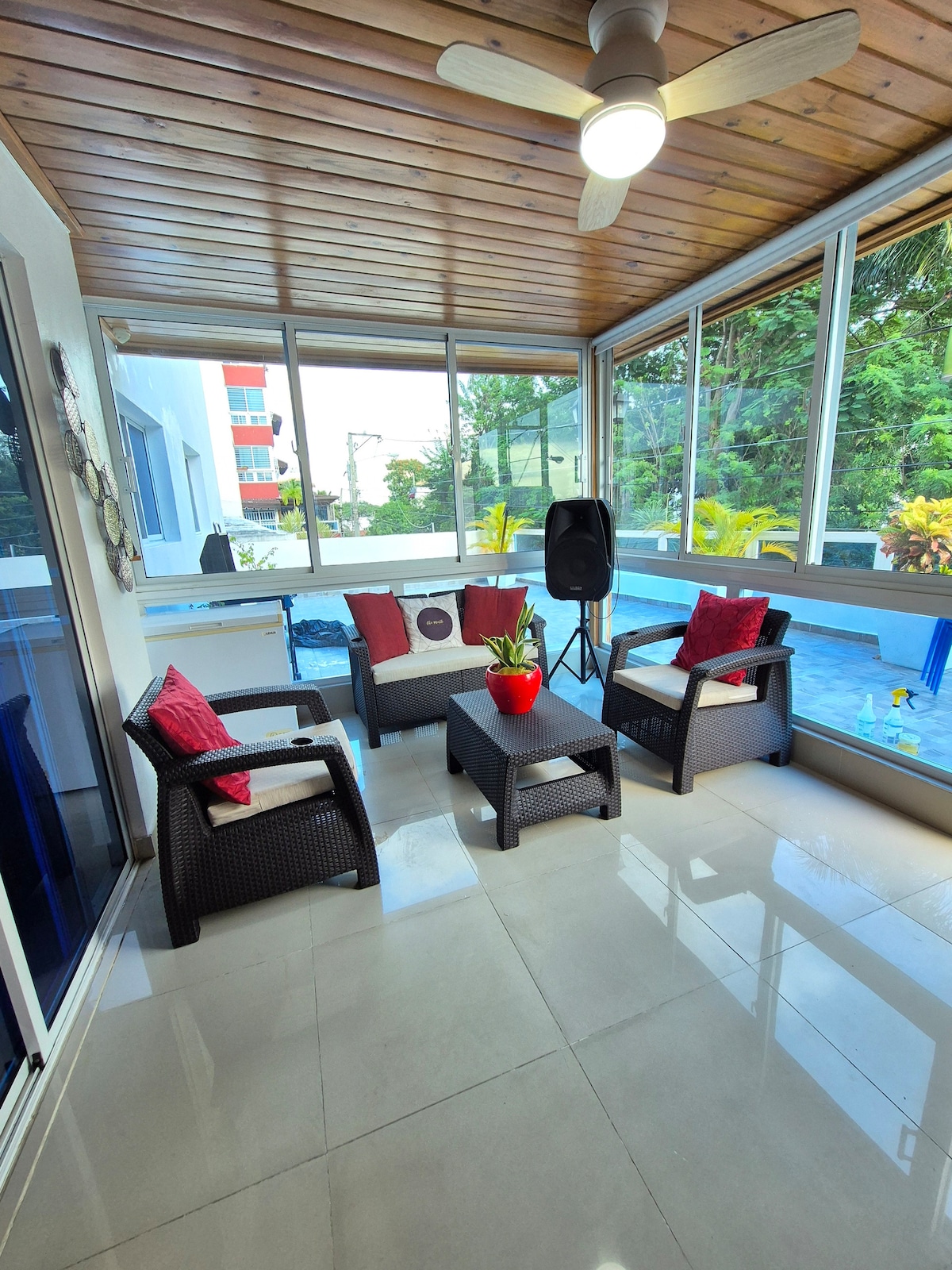 A sunlit sitting area features a glass-enclosed space with a ceiling fan, wicker furniture, and cushions in shades of red. A small coffee table is positioned at the center, accompanied by a decorative plant. The backdrop includes glimpses of greenery and a pool area visible through the glass.
