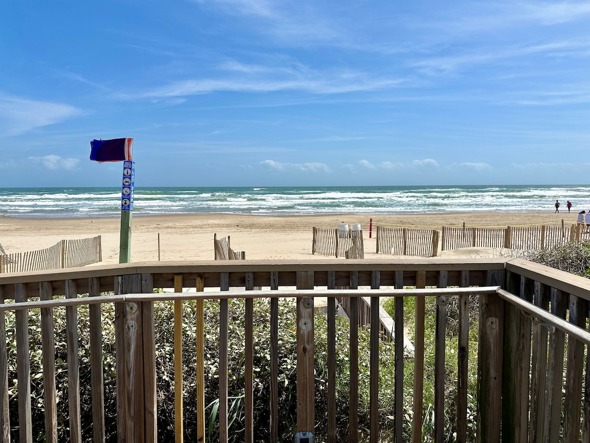 The image captures a view of the beach from a wooden balcony. Soft waves roll onto the sandy shore, bordered by a wooden fence. A colorful beach flag stands nearby, indicating beach access, while a few people stroll along the shoreline in the distance.