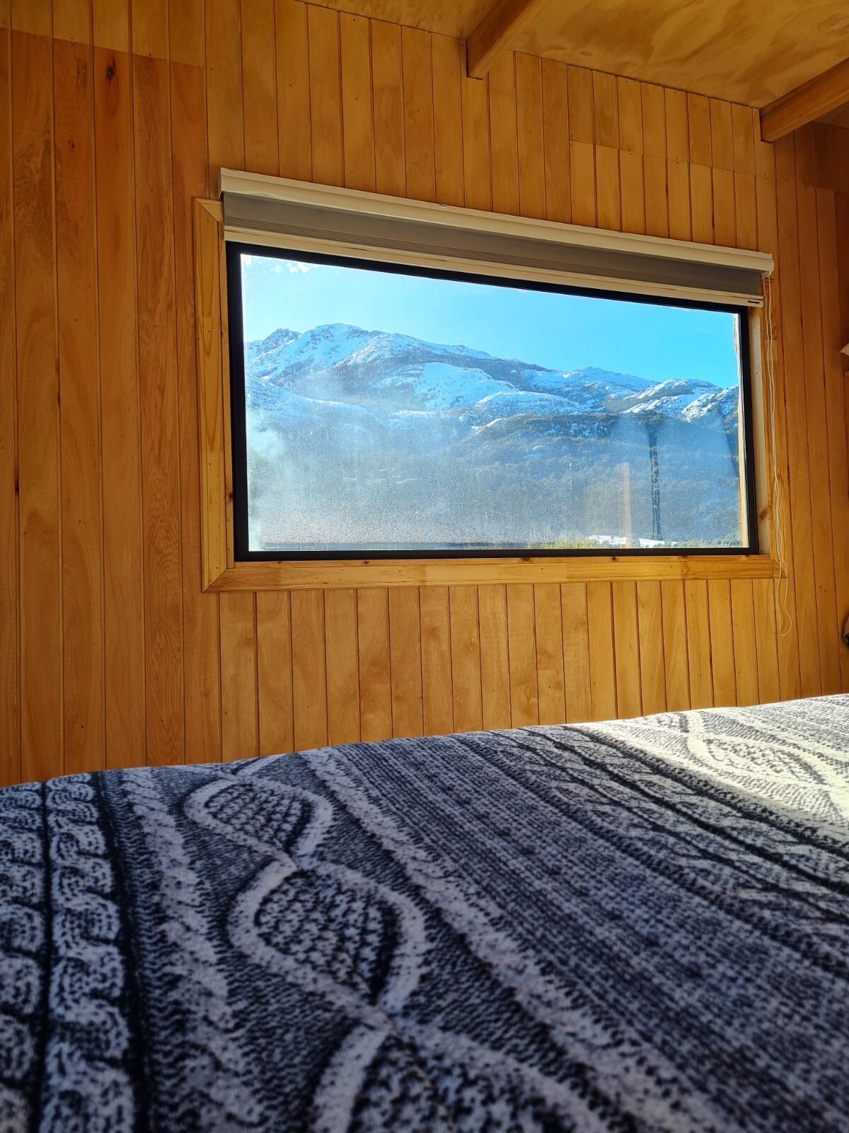 A cozy bedroom features wooden paneling and a spacious bed covered with a patterned blanket. A large window offers a view of snow-capped mountains, allowing natural light to fill the room.