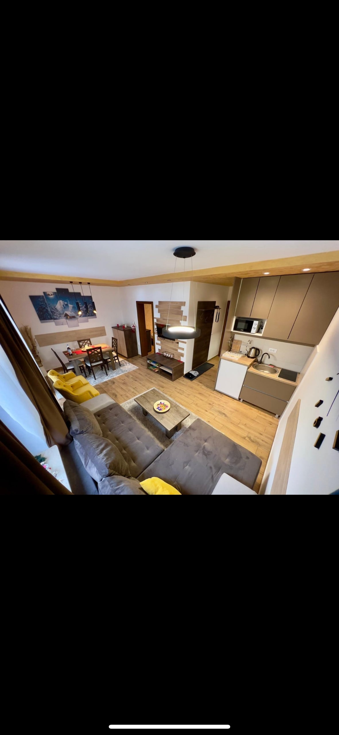 A modern living space is shown, featuring an open layout with a comfortable sofa, small dining area, and a well-equipped kitchen. Light wooden flooring enhances the area. Wall art adds a contemporary touch, while natural light is received through the nearby windows.