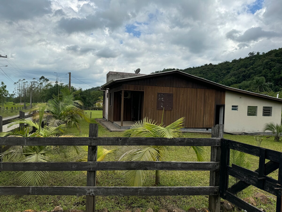 The house features a wooden exterior with large windows, surrounded by greenery and a black wooden fence. The landscape includes patches of grass and young palm trees, with rolling hills visible in the background under a cloudy sky.