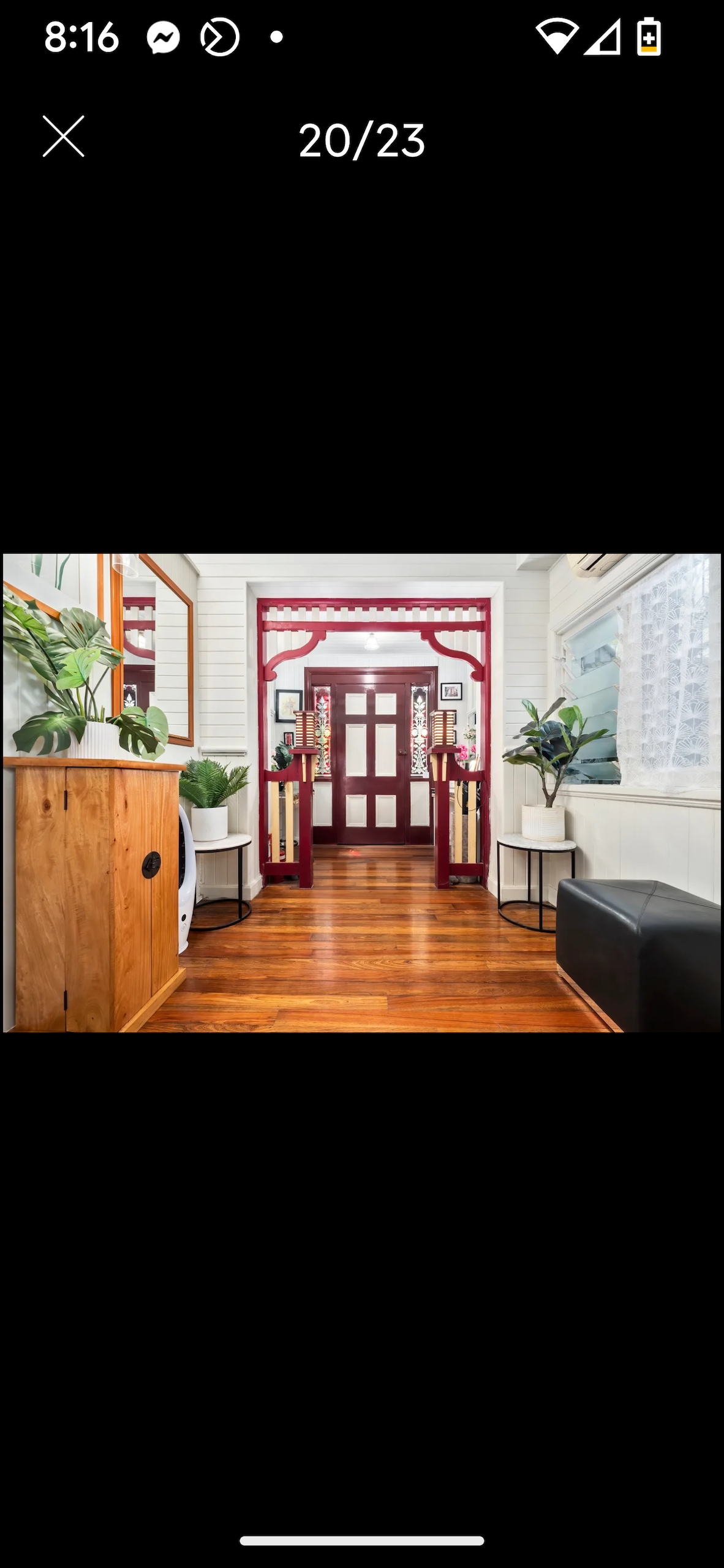 A welcoming hallway is showcased, featuring polished wooden flooring and decorative plants in planters. An ornate red archway frames an entrance bordered by two stylish folding screens. Bright light filters through the windows, highlighting the clean and inviting atmosphere of the space.