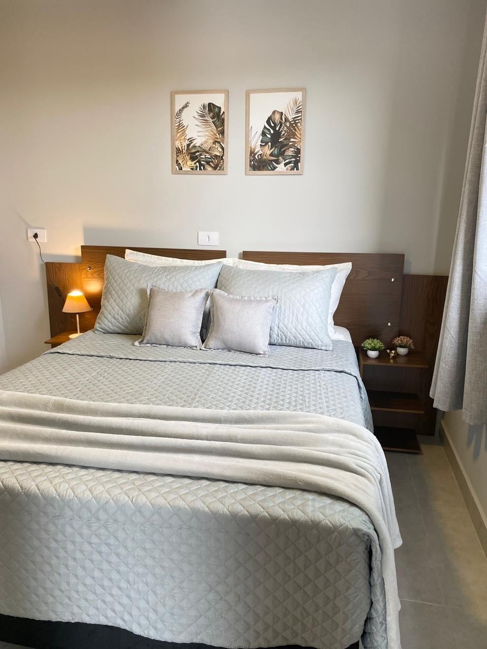 A well-organized bedroom features a spacious bed adorned with soft bedding and coordinating decorative pillows. Wall art depicting plants adds a serene touch. A bedside lamp illuminates the area, while a window covered by light curtains offers a soft, natural glow.