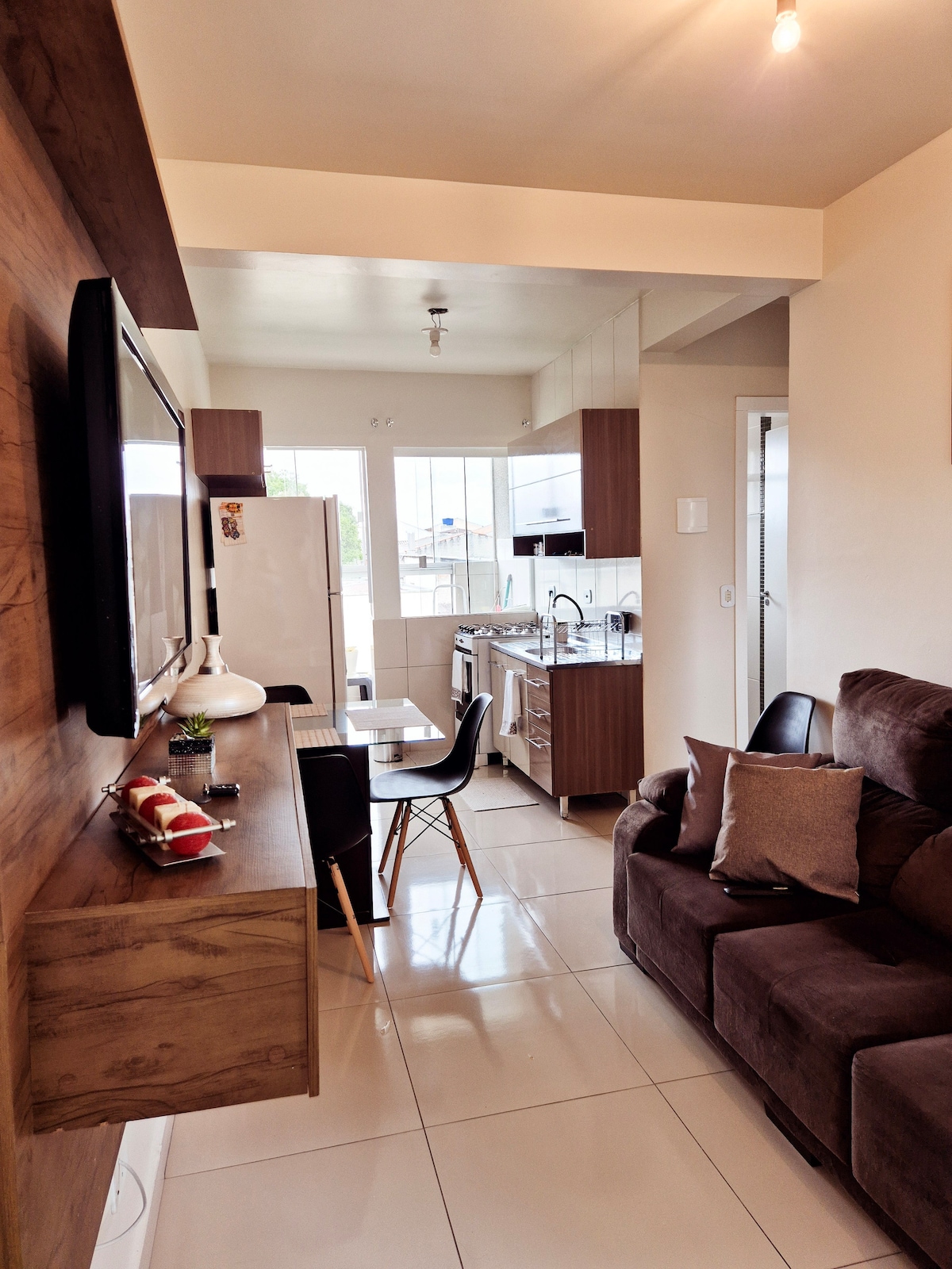 A modern living space features a dark brown sofa adorned with cushions, accompanied by a small dining area with a table and black chairs. The kitchen is visible, showcasing sleek wooden cabinetry and stainless steel appliances. Natural light fills the room through large windows.