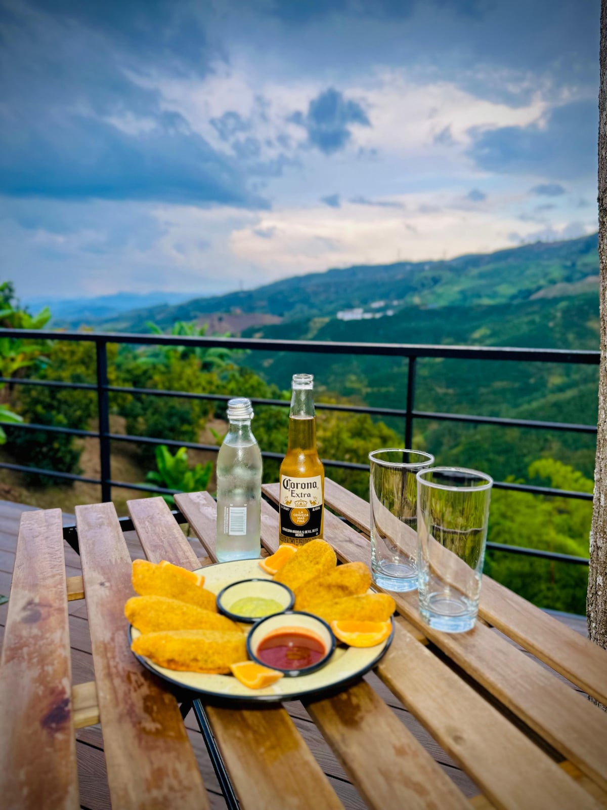 A wooden table set outdoors features a platter of tacos accompanied by salsa and slices of orange. Two bottles of beer are positioned next to sparkling water, with clear glasses arranged beside them. The background displays lush green hills and a cloudy sky.