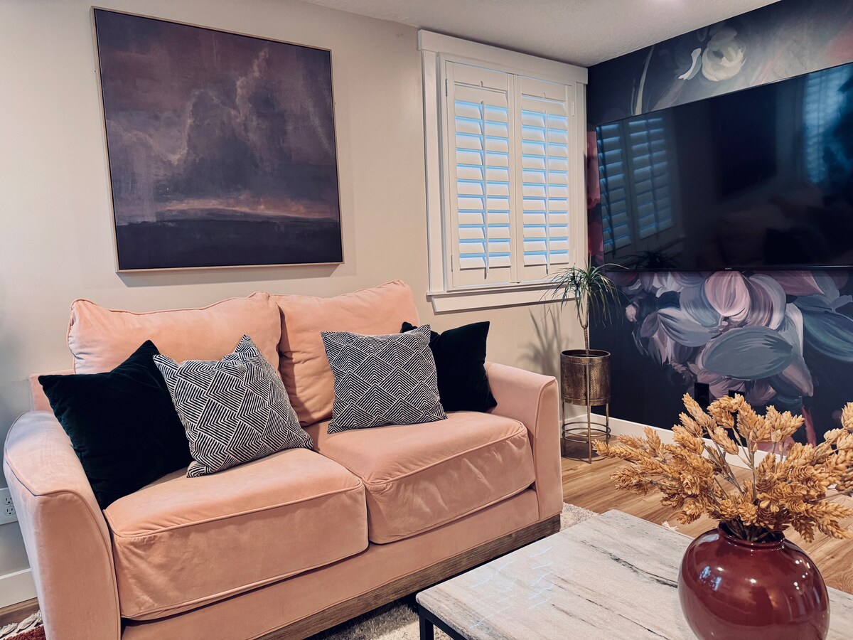 A soft pink sofa is positioned against a wall adorned with a floral mural. It is complemented by decorative pillows in various patterns. A modern coffee table sits in front, with a vase of dried flowers adding a natural touch. Light filters through the window with plantation shutters.