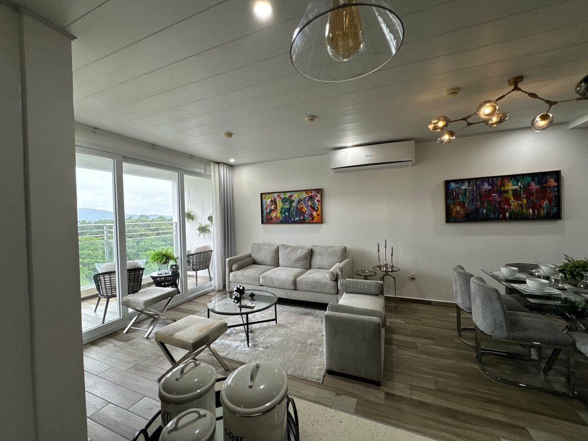 A bright living area features large glass doors leading to a balcony, providing outdoor views. A comfortable gray sofa and decorative chairs surround a coffee table. Artwork adorns the walls, and a dining table is set nearby, contributing to a welcoming ambiance.