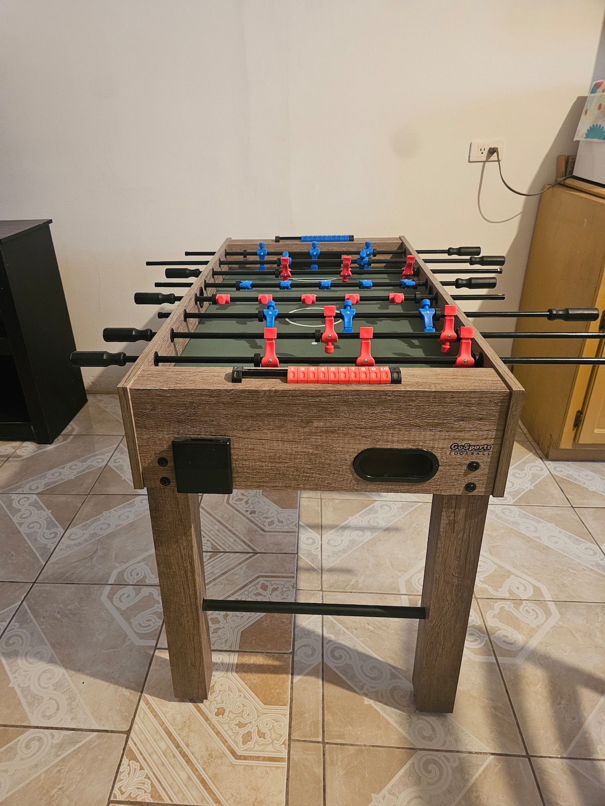 A foosball table is positioned centrally, featuring red and blue player figures on each side. The table's wooden frame is complemented by a flat green playing surface. Surrounding the table, a patterned tile floor adds visual interest to the space.