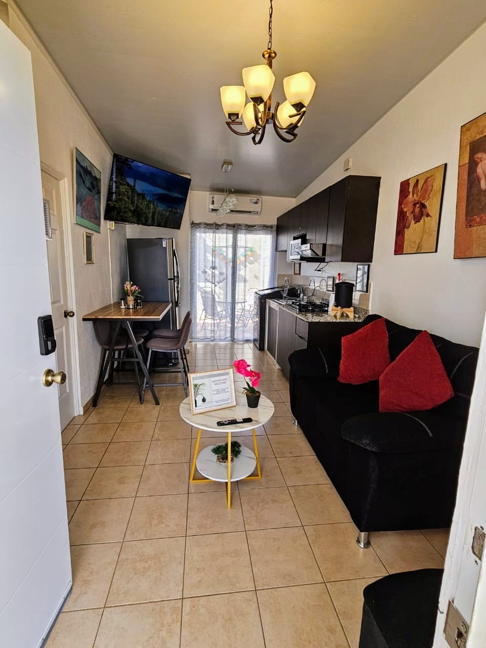 A compact living space is shown, featuring a small dining table with high chairs and a cozy sofa adorned with colorful pillows. A wall-mounted TV is installed above the kitchen area, which includes cabinetry and appliances. Natural light streams in through sliding doors leading to a balcony.