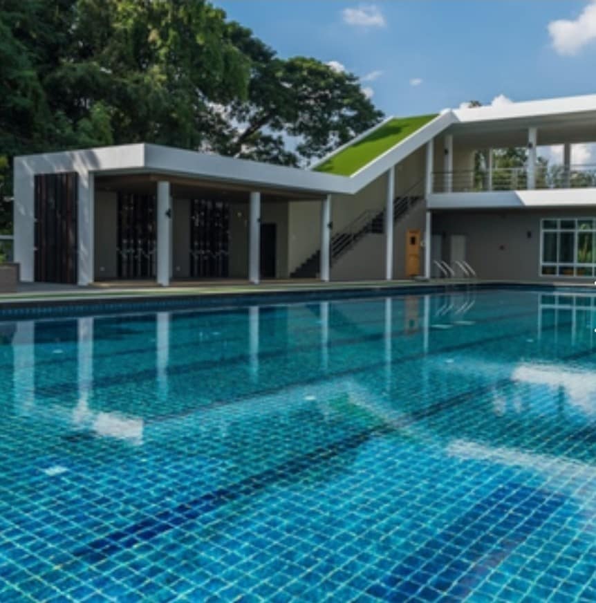 A modern public pool is surrounded by a contemporary building. The pool's clear blue water reflects the sky and greenery, with lounge areas visible beyond. A staircase leads up to the upper level of the structure, providing access to additional amenities.