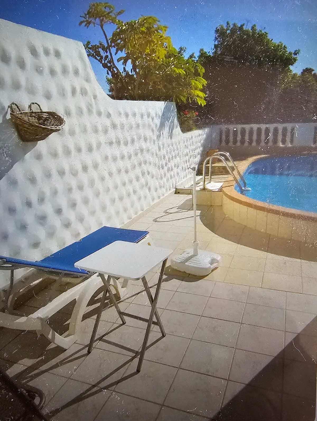 A private outdoor area is shown, featuring a pool with a gently curved edge. A small, blue table is positioned beside a lounge chair. A light-colored wall with a textured surface surrounds the space, complemented by a small plant and a basket fixed on the wall.