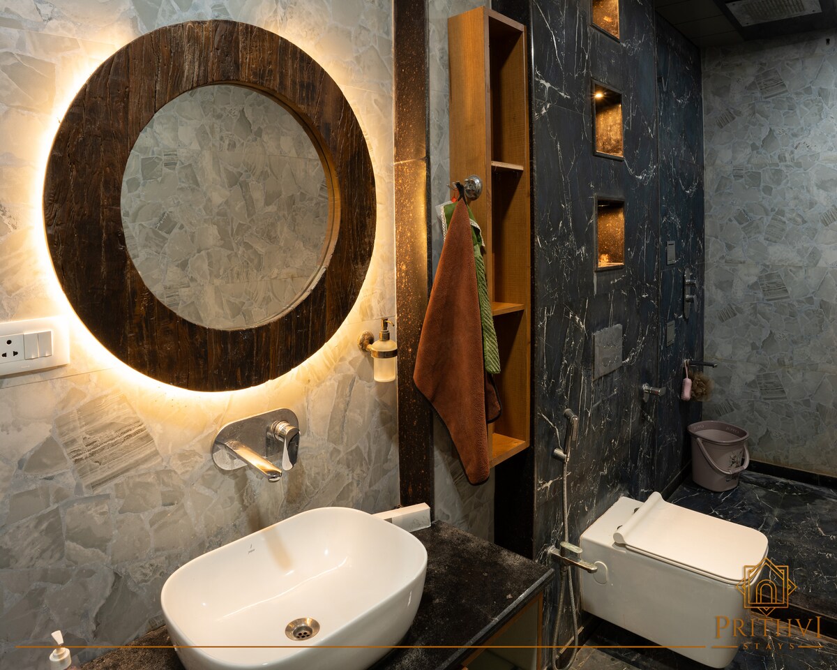 The stylish bathroom features a round mirror framed with warm lighting above a sleek sink. A wooden shelf displays neatly arranged toiletries, while a modern toilet is positioned beside a dark stone wall. The overall design combines luxury with functionality.