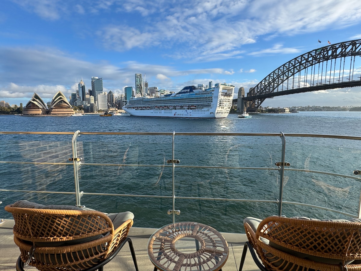 A balcony view features two wicker chairs positioned beside a round table, overlooking the water. The iconic Sydney Harbour Bridge and Opera House are visible in the background, with a cruise ship passing through the harbor under a bright blue sky.