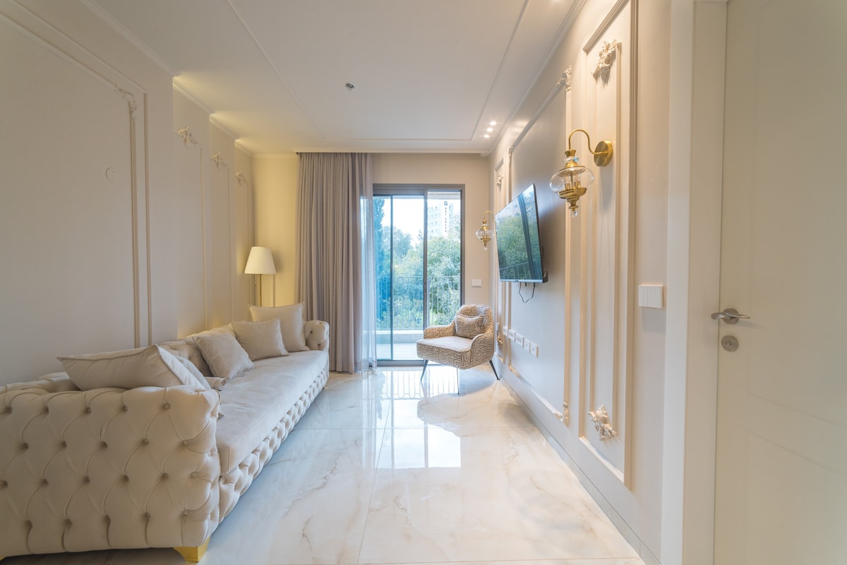 A spacious living room is shown with a light-colored sofa, elegantly tufted, arranged against a wall. Natural light enters through large glass doors leading to a balcony, and decorative wall fixtures add a touch of sophistication.