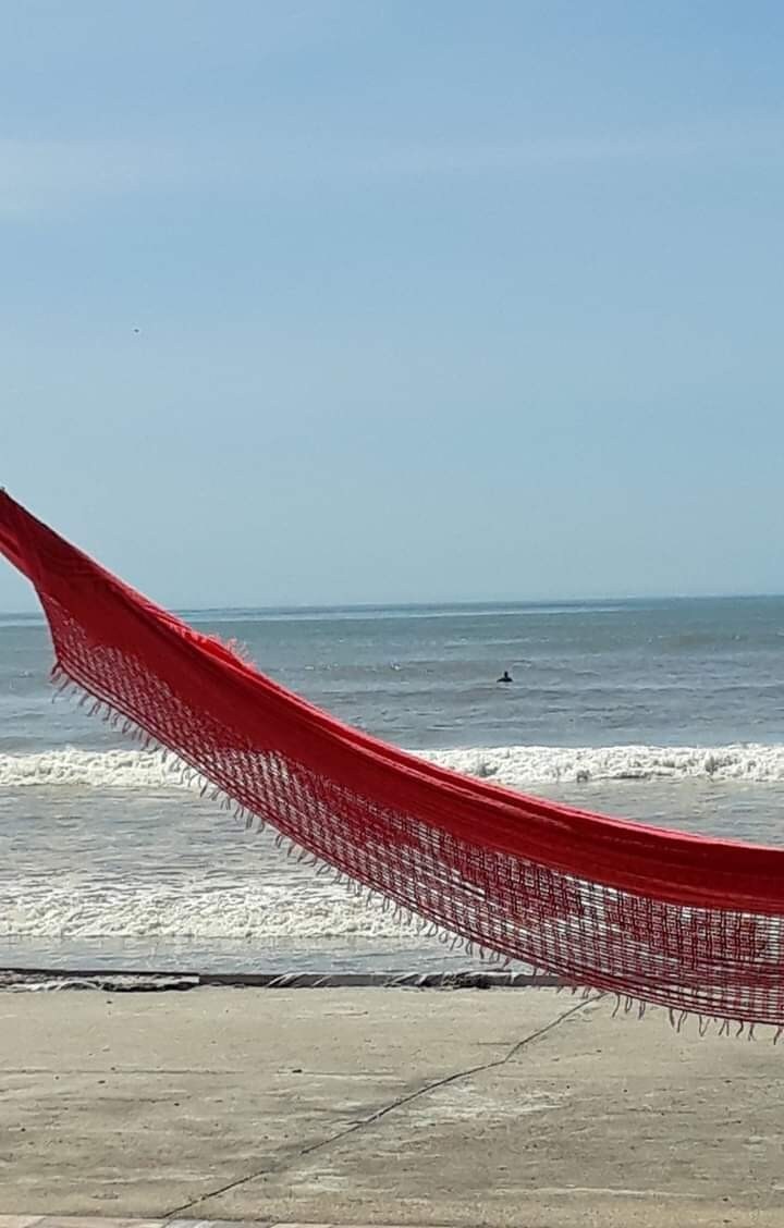 A red hammock is suspended between posts, offering a relaxing view of the ocean. Gentle waves are visible in the distance, with a person surfing further out. The sky is clear with a few clouds, complementing the serene beach atmosphere.