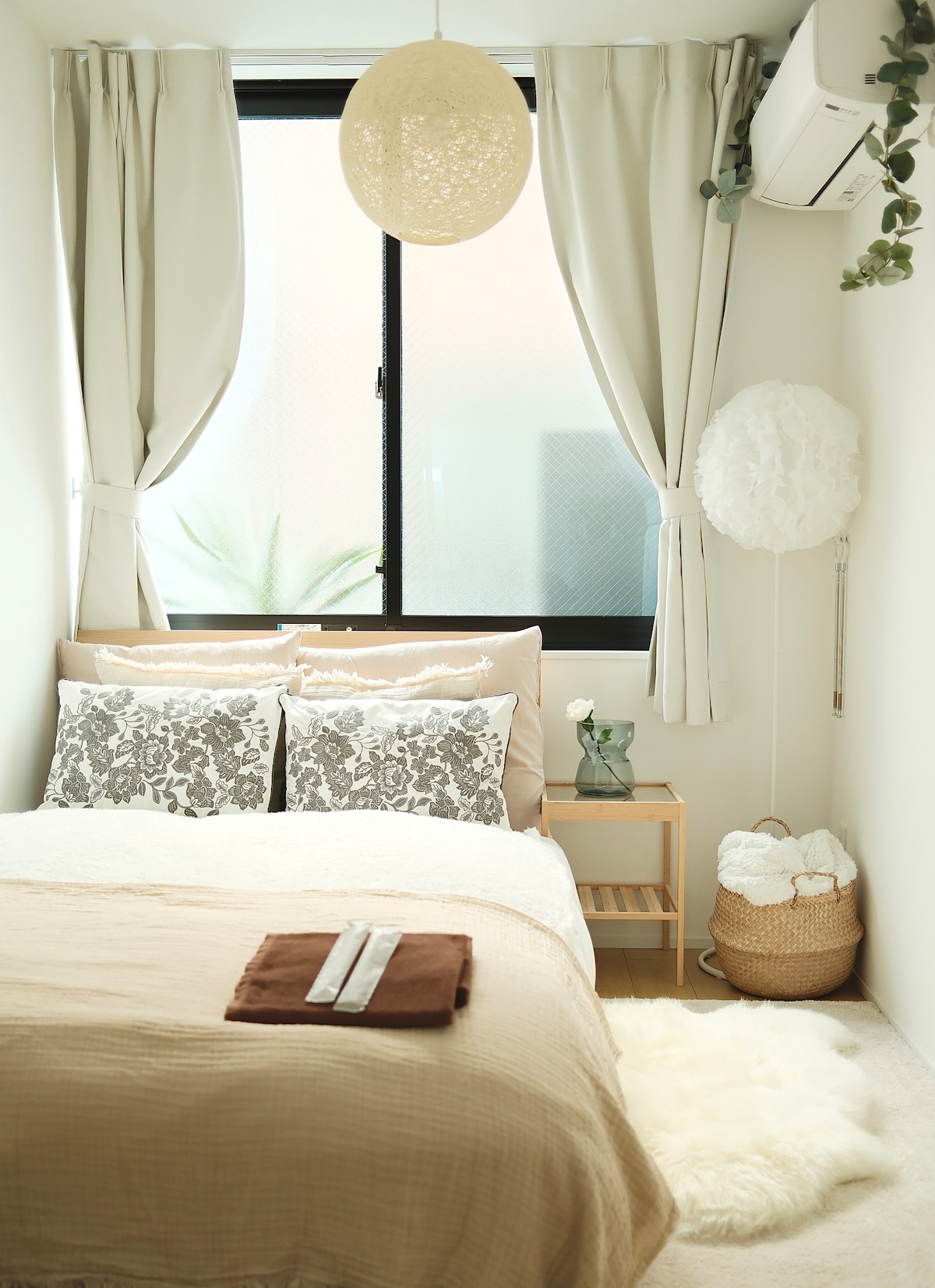 A calming bedroom features a spacious bed adorned with soft bedding and decorative pillows. Natural light filters through sheer curtains, illuminating a nightstand with a vase. Cozy textiles, including a light rug, and a hanging light add to the serene ambiance of the room.
