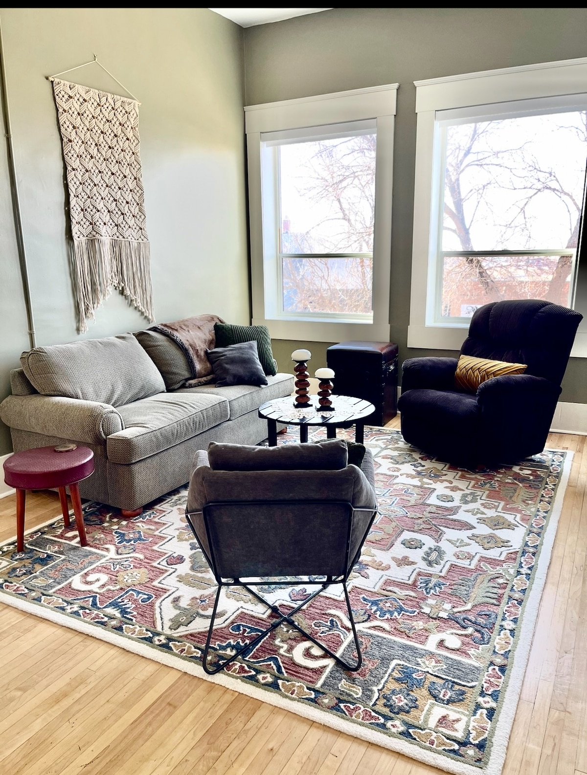 A comfortable living area features a gray sofa with decorative pillows, a patterned area rug, and a small round table. A black armchair adds contrast, while two large windows allow natural light to illuminate the space, offering views of the surroundings.