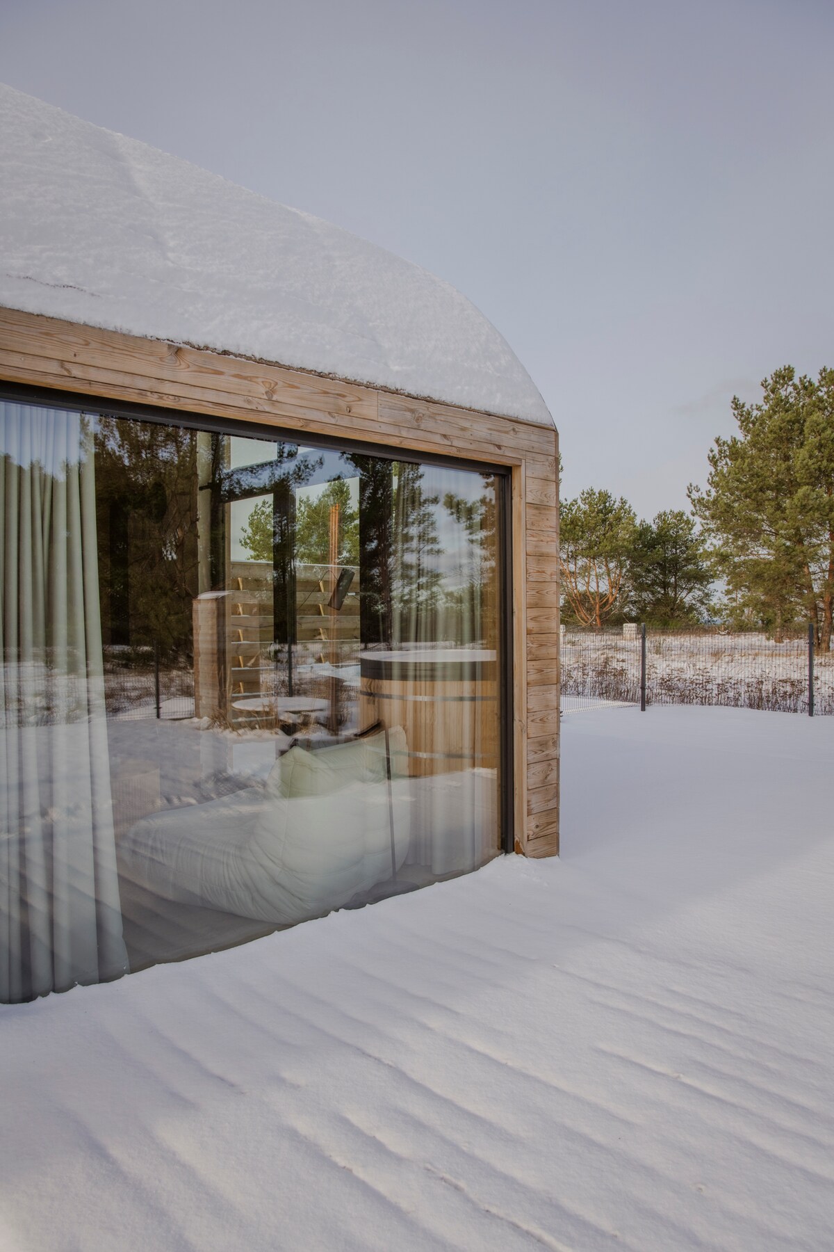 A cozy cabin is surrounded by snow-covered ground, featuring large glass windows that provide views of the natural landscape. The structure has wooden accents and a curved roof, creating a harmonious blend with the serene, wintry environment.