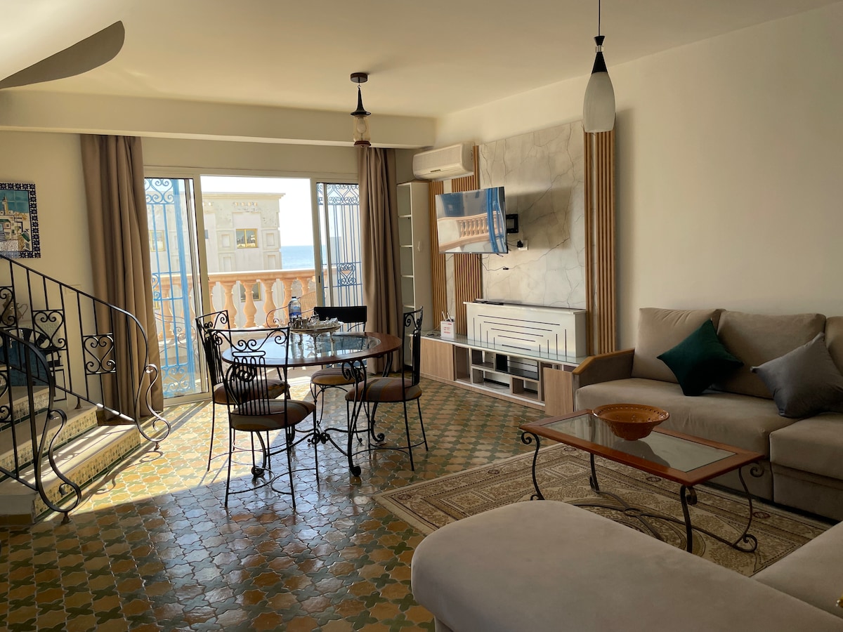 A spacious living area features a large sofa and a coffee table, complemented by dining seating for four. Natural light floods the space through glass doors, leading to a balcony with a sea view. The tiled floor adds character, alongside decorative elements in a modern style.