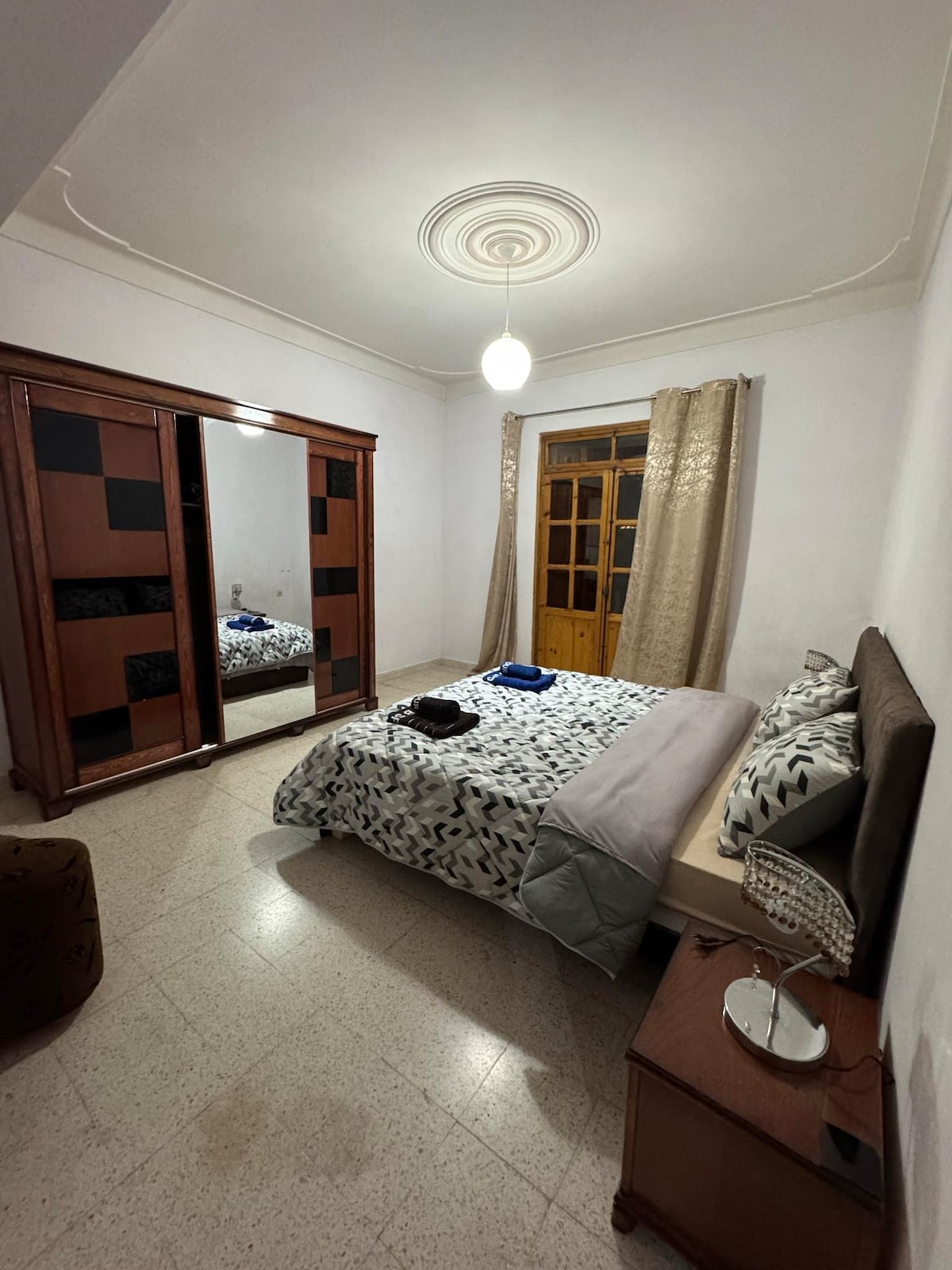 A spacious bedroom features a large bed dressed with a patterned duvet. A wooden wardrobe with mirrored panels stands against one wall, reflecting natural light from the window. Tidy bedside tables hold lamps, and curtains gently frame the window, enhancing the serene atmosphere.