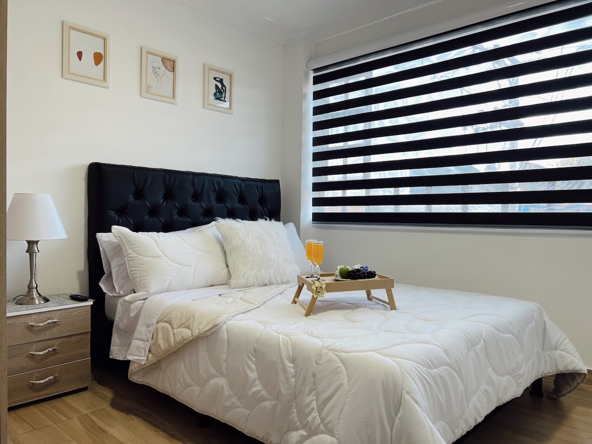 A spacious bed with a tufted black headboard occupies the center of the room. Soft white bedding and decorative pillows complement the decor. A bedside lamp and a small nightstand are positioned nearby. The window, covered with dual-layered blinds, allows for soft natural light.