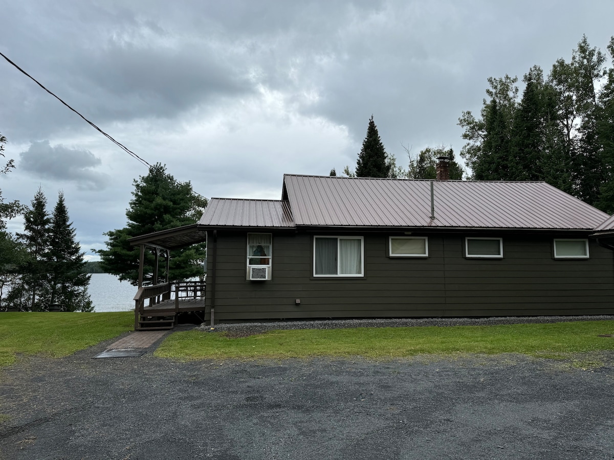A rustic cabin featuring a metal roof and dark wooden siding is set against a backdrop of lush trees. The porch leads to an accessible entryway, while views of Mud Lake are visible beyond the grassy yard. Cloudy skies add to the serene atmosphere.