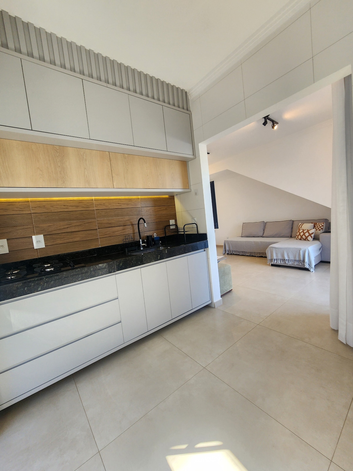 A modern kitchen area features sleek cabinetry in light tones, complemented by a dark granite countertop. Beyond, a cozy living space is visible, showcasing a light grey sofa with decorative cushions. Natural light filters in through a nearby window, illuminating the open layout.