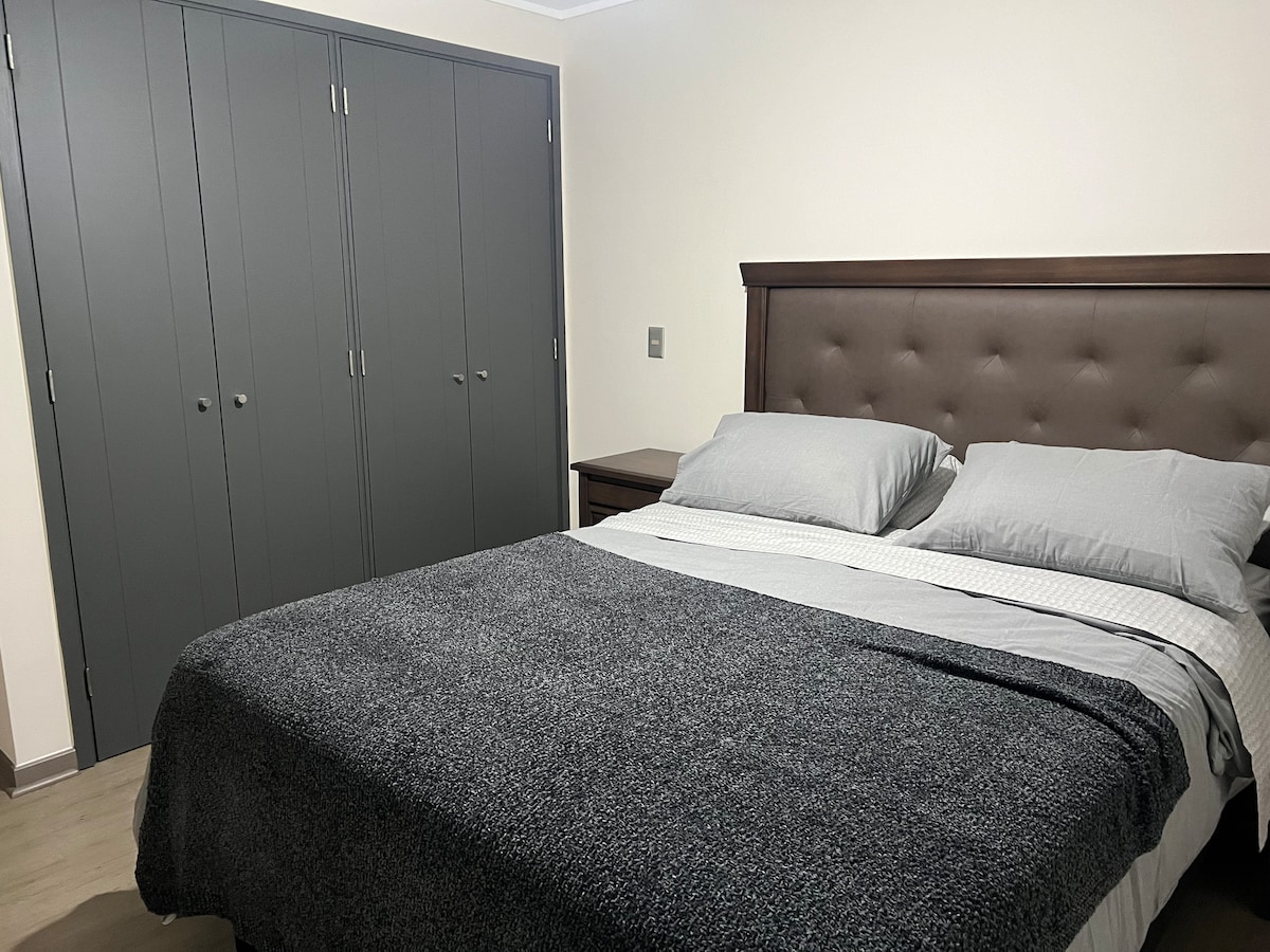 A well-appointed bedroom features a spacious bed with neutral-colored linens. Soft pillows rest against a tufted headboard. A sleek nightstand and a built-in wardrobe are visible, providing ample storage options. The room is brightly lit, creating a warm and welcoming environment.