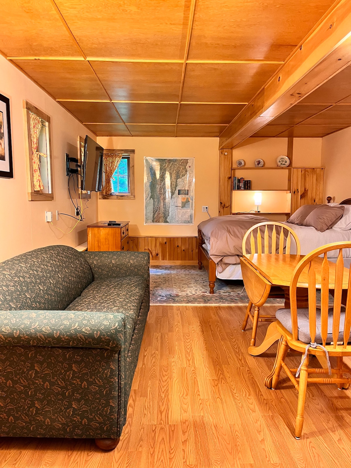 A cozy studio apartment is showcased, featuring a spacious bed elevated on a wooden platform. A dining table with wooden chairs is positioned by a wall, while a green sofa adds seating. A television is mounted on the wall, and natural light enters through a nearby window.