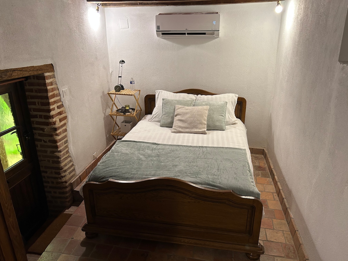 A cozy bedroom features a spacious bed with light bedding and decorative pillows. A wooden headboard complements the rustic charm. An air conditioning unit is mounted on the wall, and a small bedside table holds a lamp. A door leads to the outside.