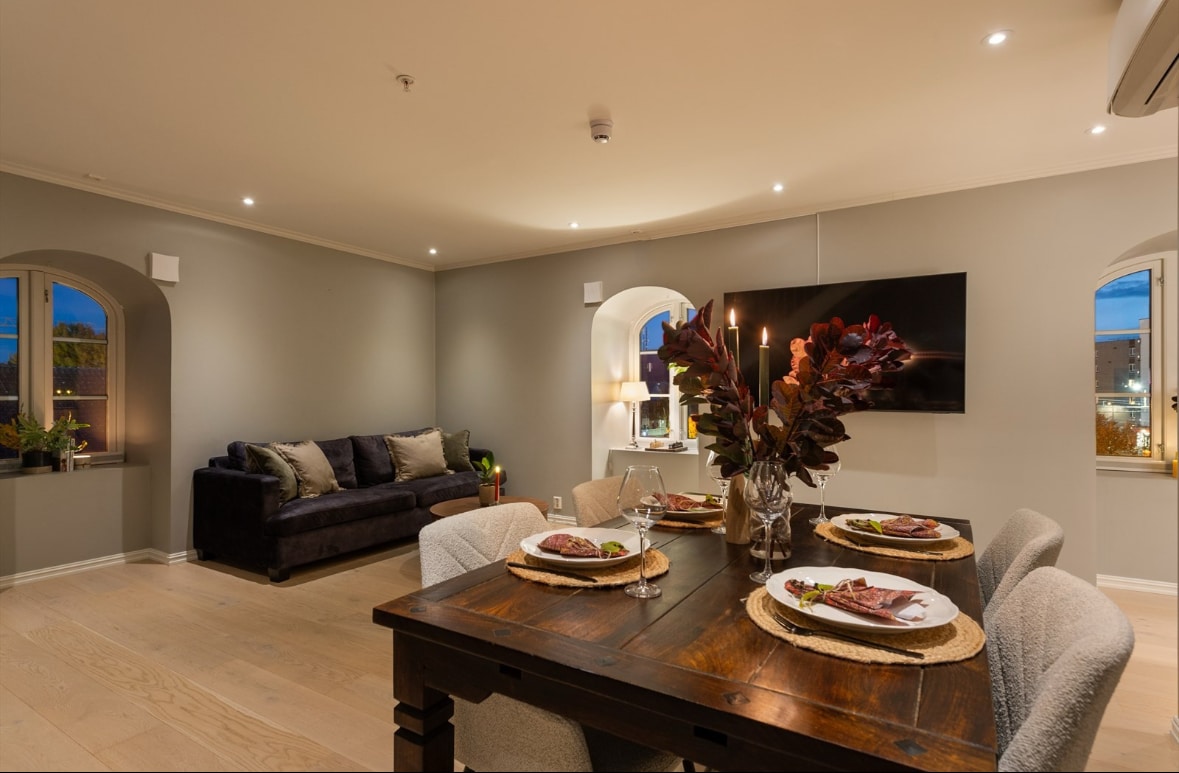 A dining area is featured, showcasing a wooden table set with plates and elegant décor. Soft lighting highlights the cozy couch situated next to an arched window, while decorative plants add a touch of nature to the space.